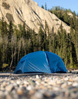 Blue tent set up on a rocky ground with trees and mountains in the background