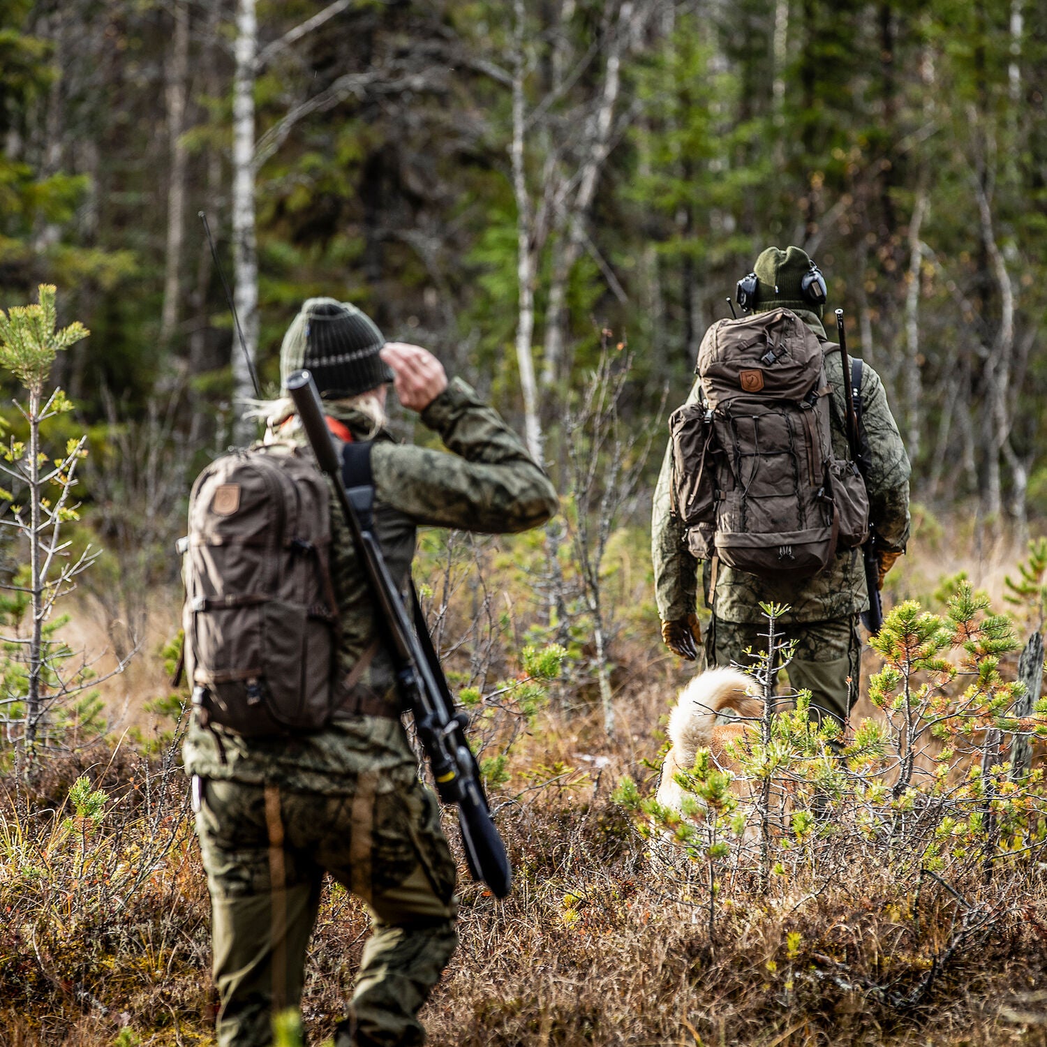 Two hunters with backpacks and rifles walking through a forest.