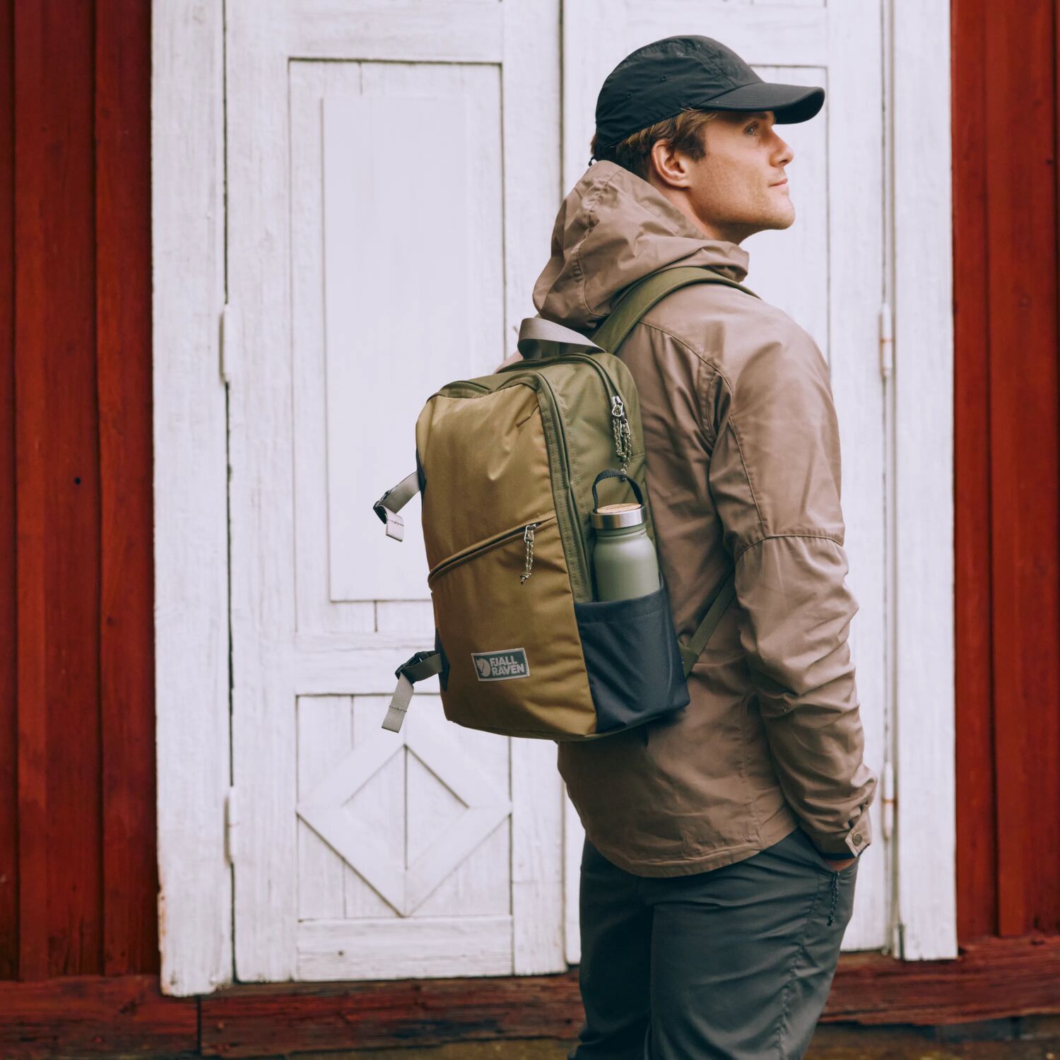 Man wearing a brown jacket and clay backpack with a red wooden door in the background