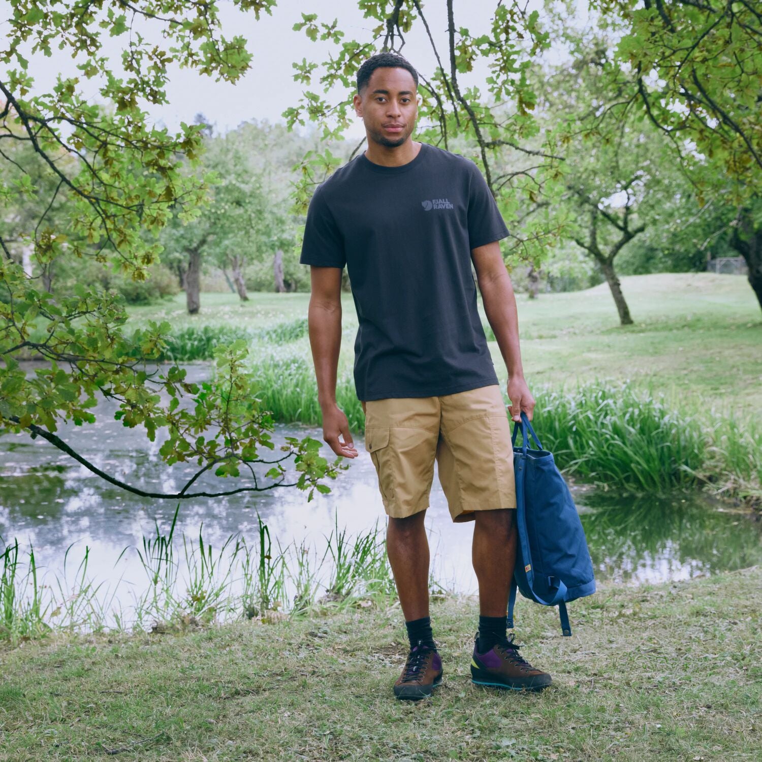 Man standing outdoors by a pond holding a blue bag, wearing a black t-shirt and beige shorts.