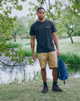 Man standing outdoors by a pond holding a blue bag, wearing a black t-shirt and beige shorts.