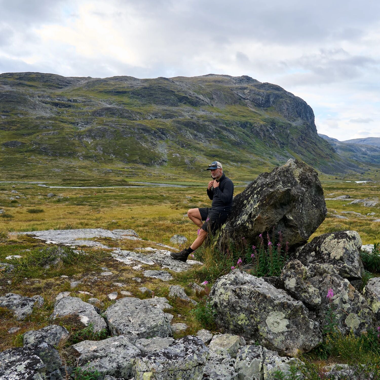 Person sitting on a rock in a mountainous landscape with grassy hills and a cloudy sky.