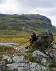 Person sitting on a rock in a mountainous landscape with grassy hills and a cloudy sky.