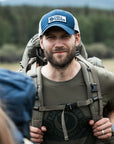 Man wearing a blue cap and carrying a backpack with Fjällräven  branding in a forest setting.