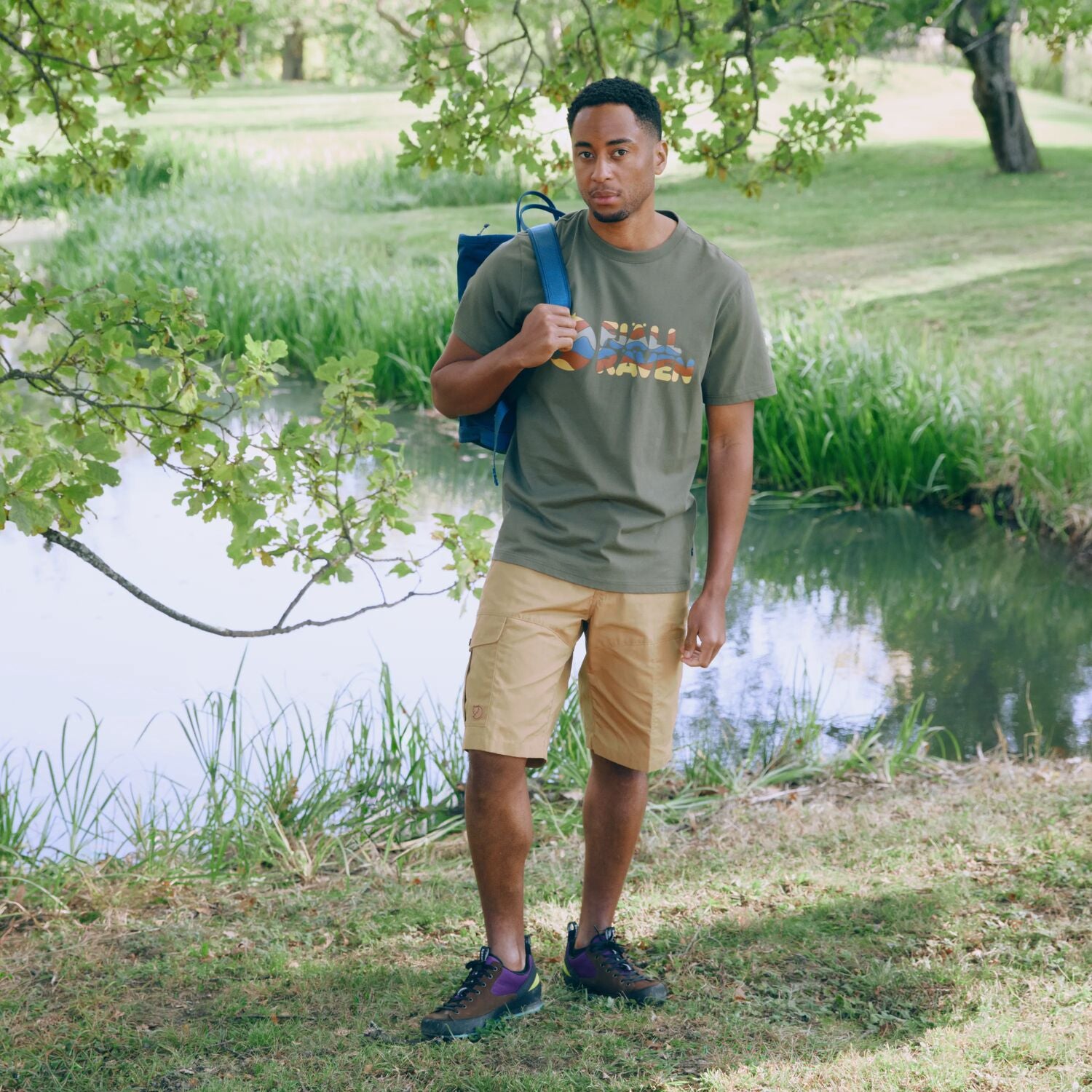 Man standing outdoors by a pond holding a blue backpack