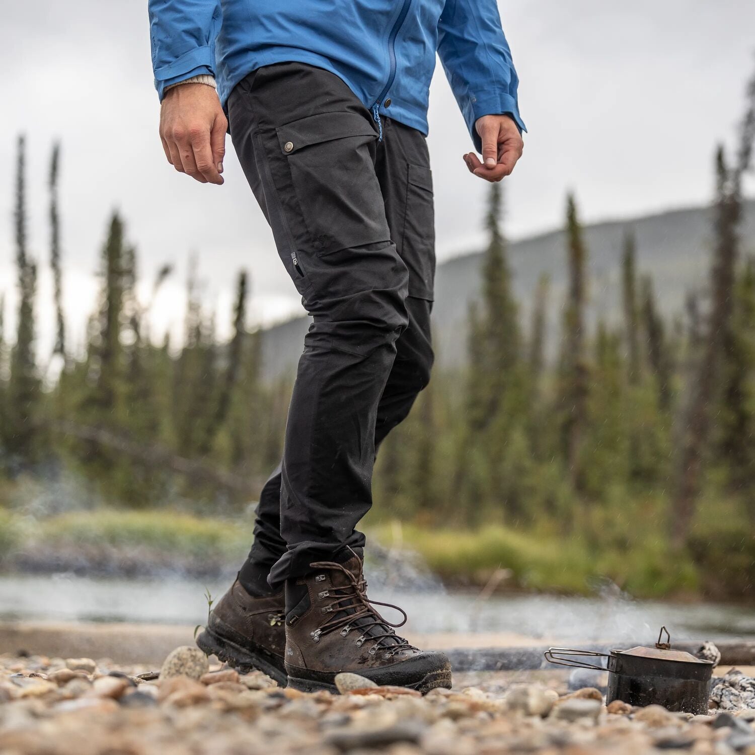 Person in outdoor setting wearing a blue jacket and black pants with hiking boots near a campfire.