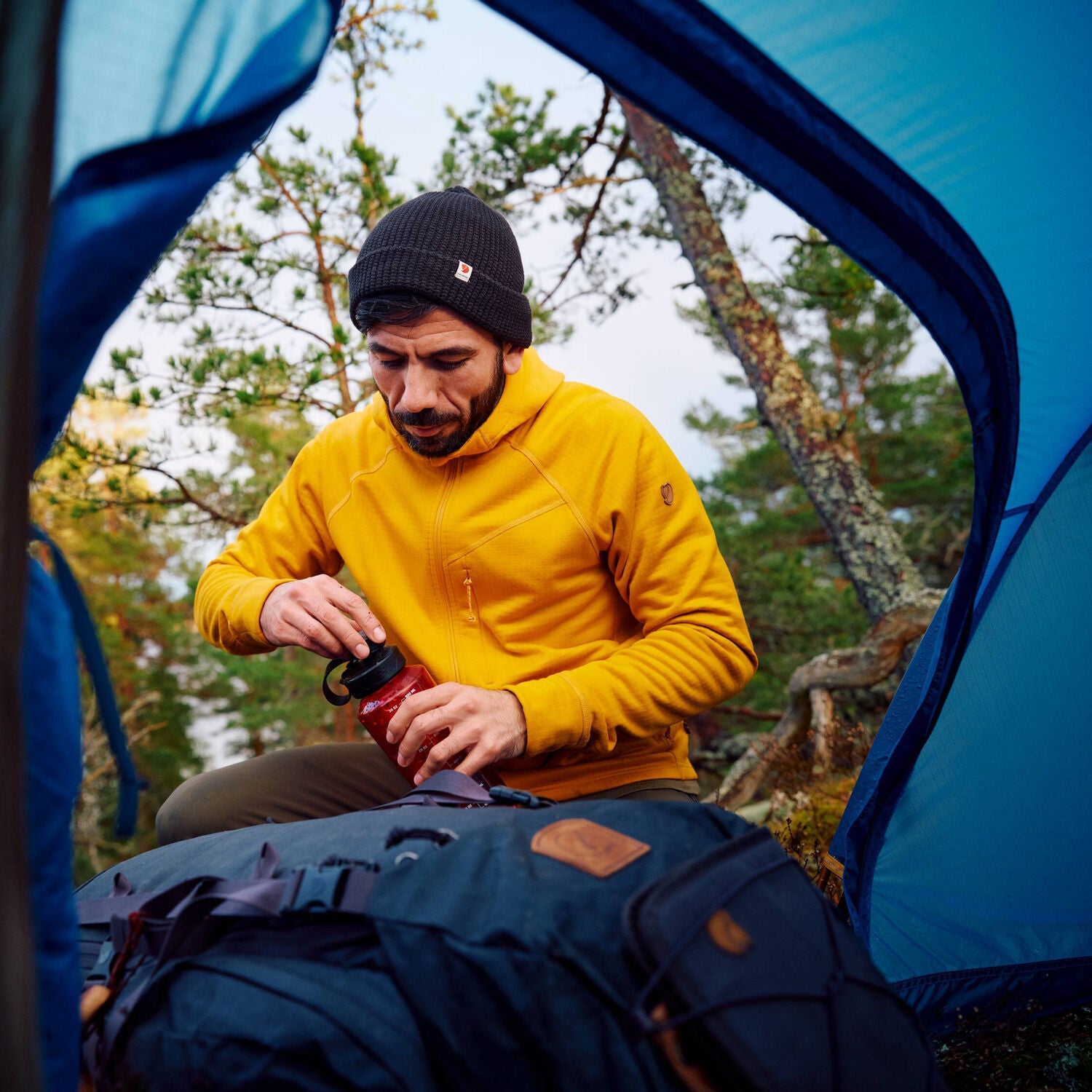 man in mustard yellow fleece jacket with hood drinking water in a tent