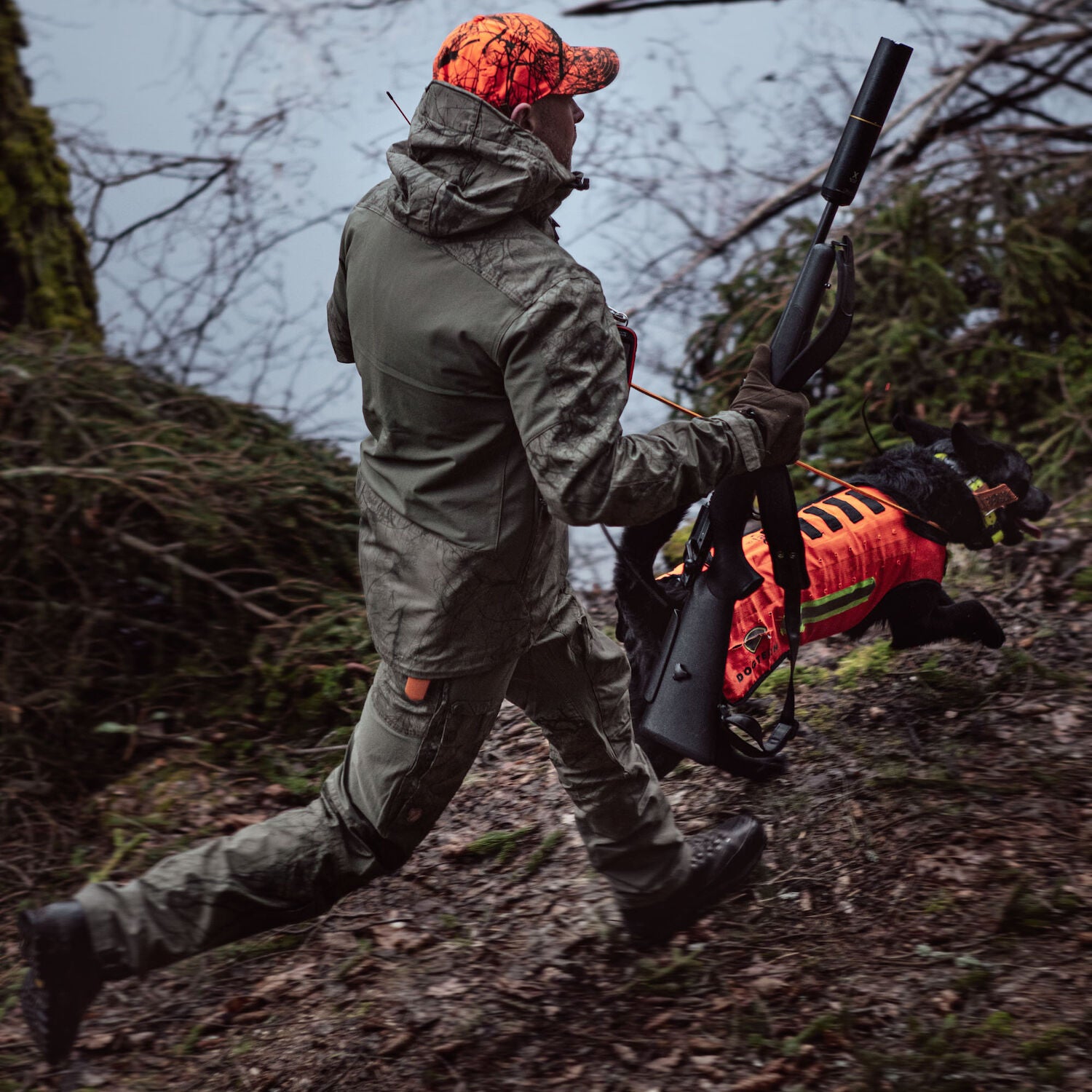 Person in hunting gear walking through a forest with a rifle and dog.