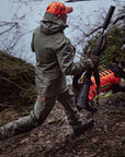 Person in hunting gear walking through a forest with a rifle and dog.