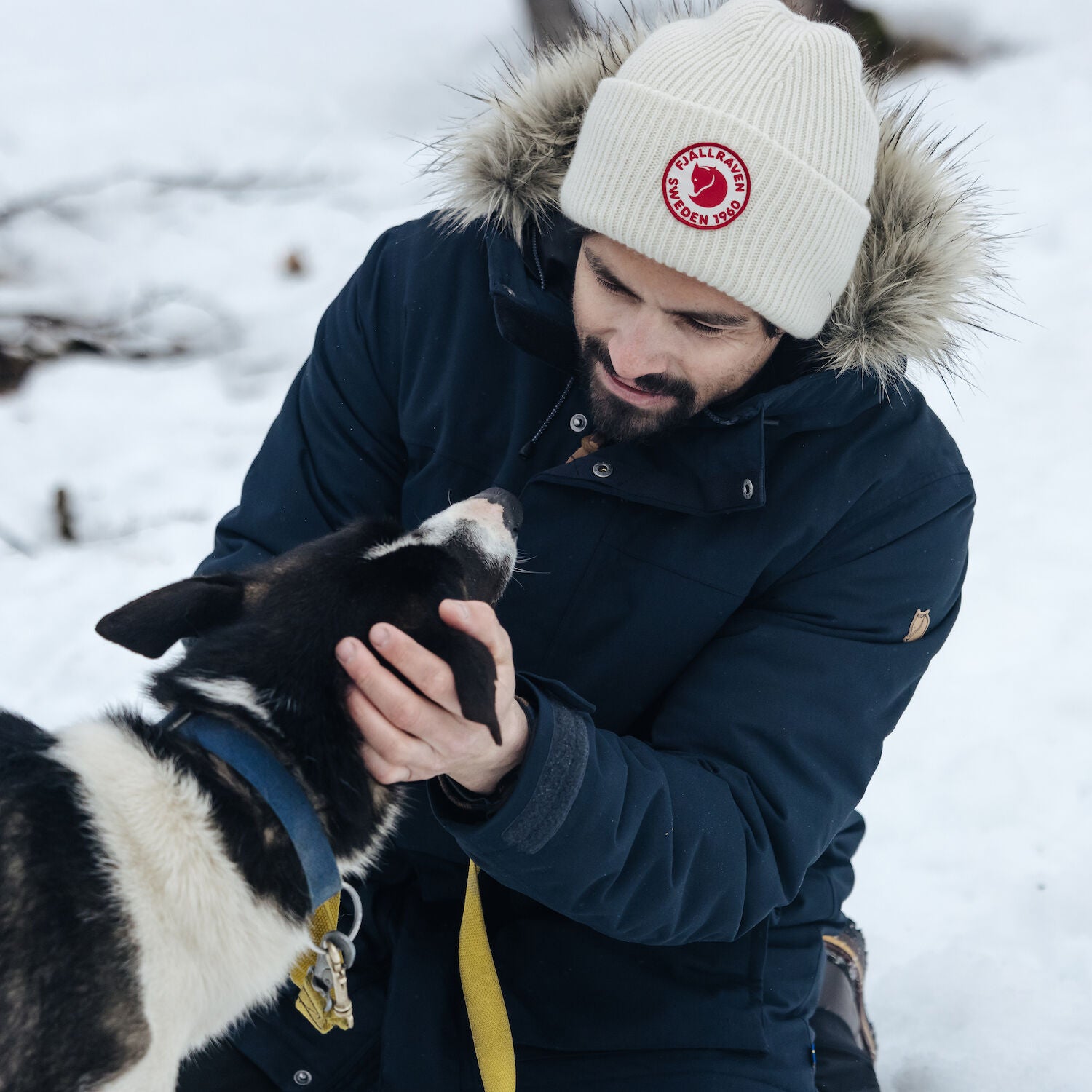 man wearing navy parka with fur hood and a white beanie playing with his dog