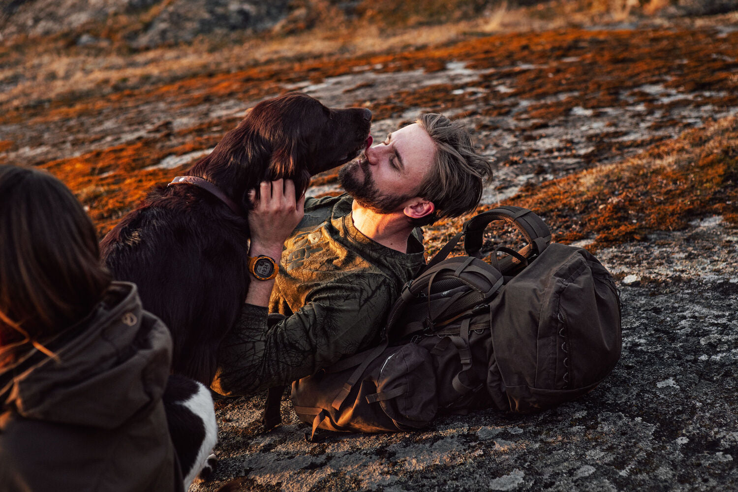 man lying on his trekking backpack on mountain surface kissing his dog