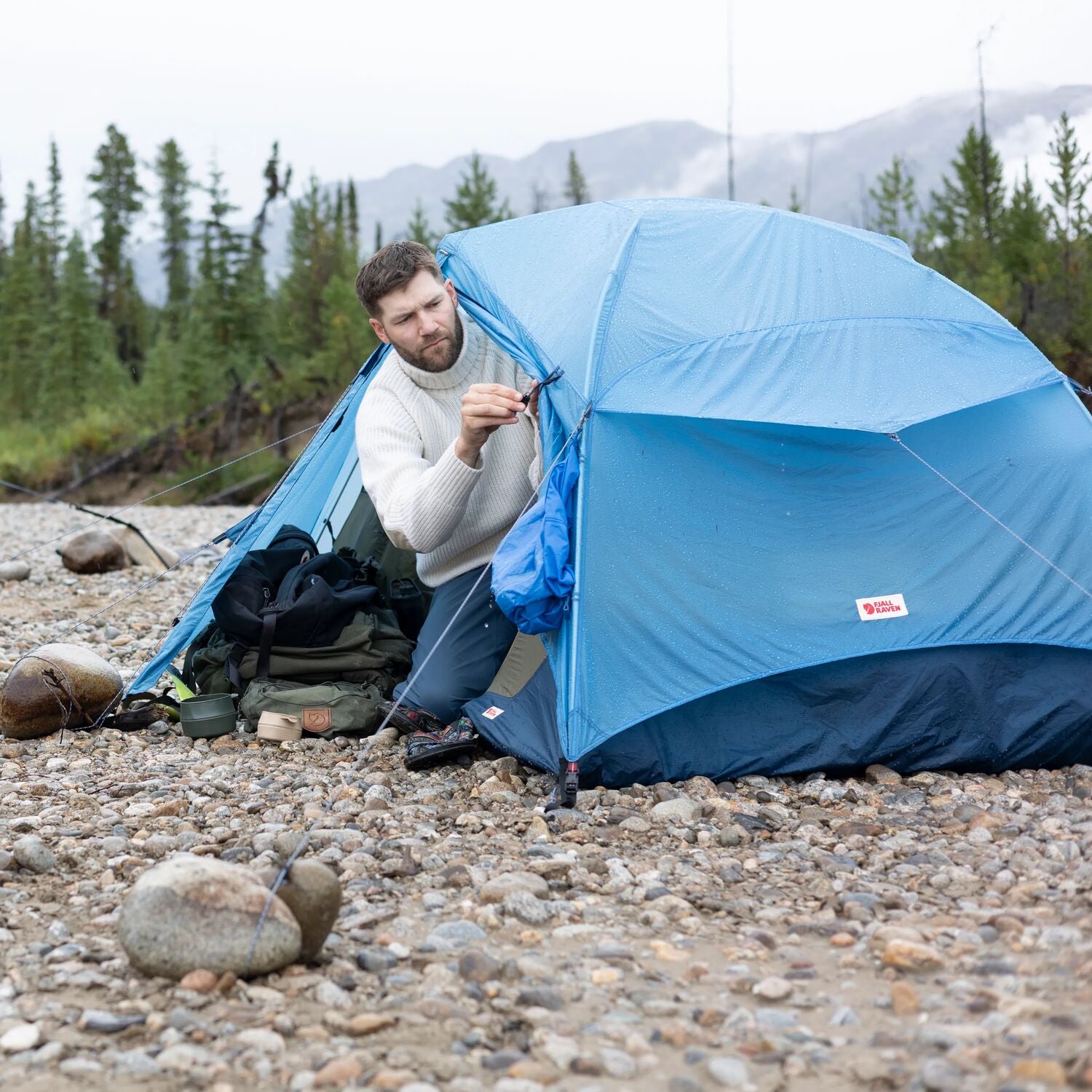 Man setting up a blue tent in a natural outdoor setting with mountains and trees in the background.