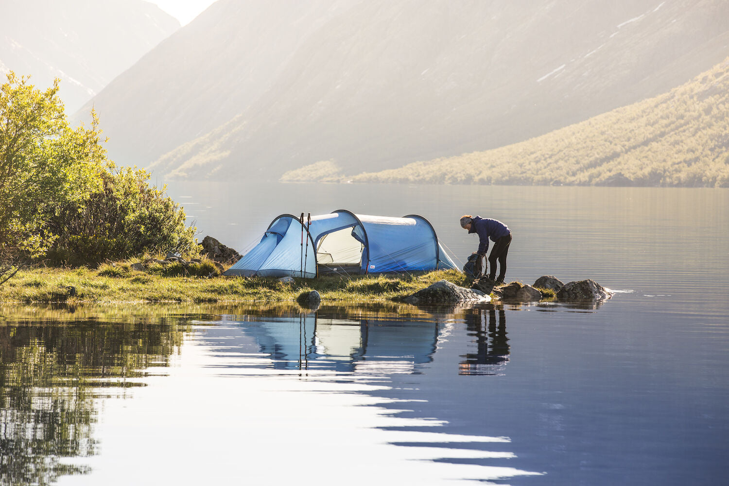 Person setting up a tent by a lake with mountains in the background