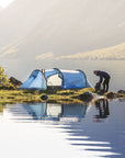 Person setting up a tent by a lake with mountains in the background