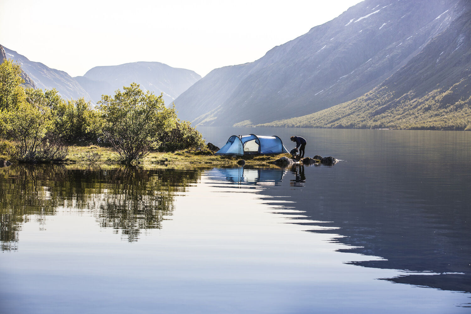 Person setting up a blue tent by a lake with mountains in the background