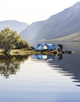 Person setting up a blue tent by a lake with mountains in the background