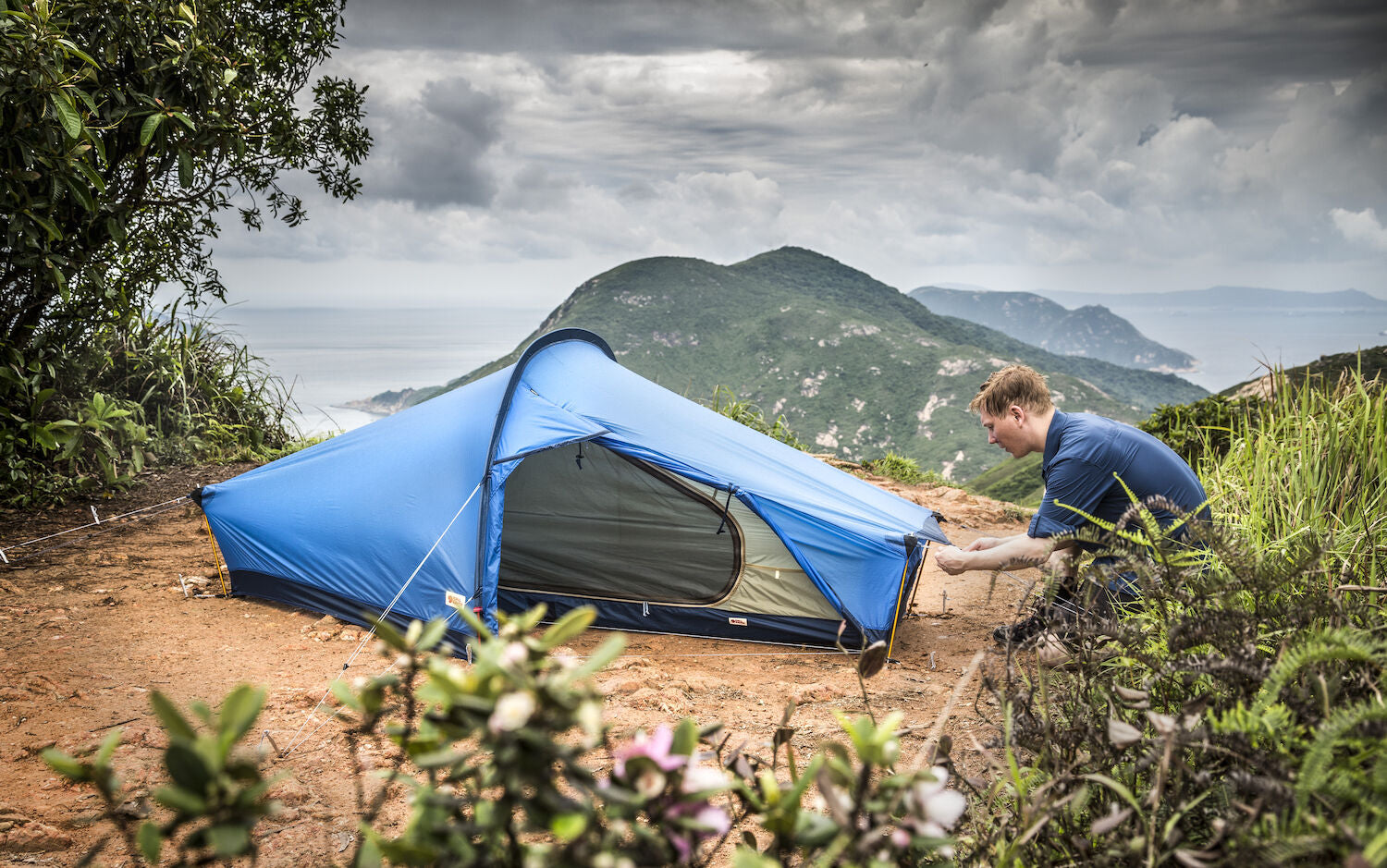 Person setting up a blue tent in a mountainous area with a scenic view.