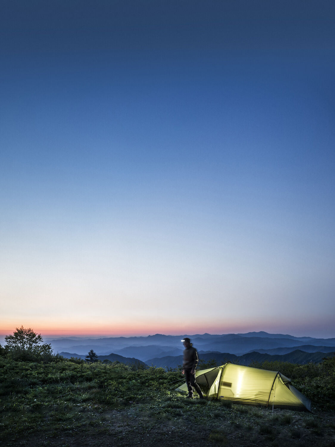 Person standing next to a tent on a mountain at dusk with a clear sky.