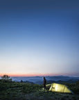 Person standing next to a tent on a mountain at dusk with a clear sky.