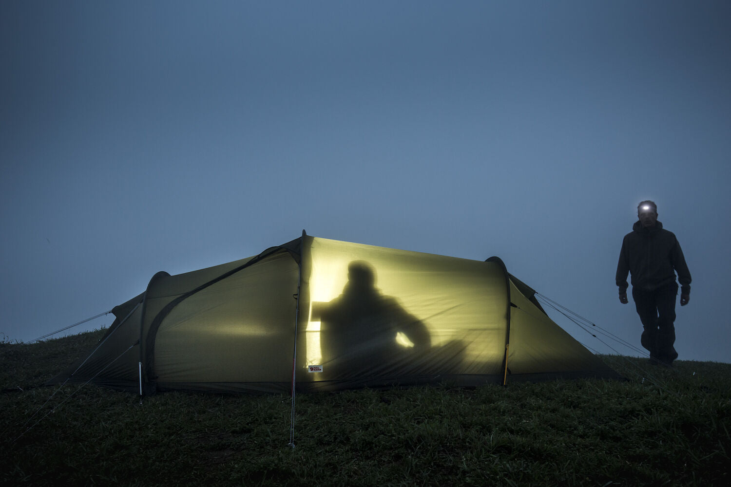 Person walking towards a illuminated tent on a grassy hill at night.