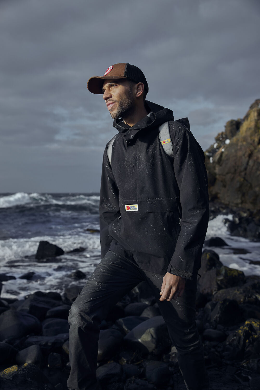man wearing raincoat jacket with Fjällräven logo carrying a backpack and brown cap
