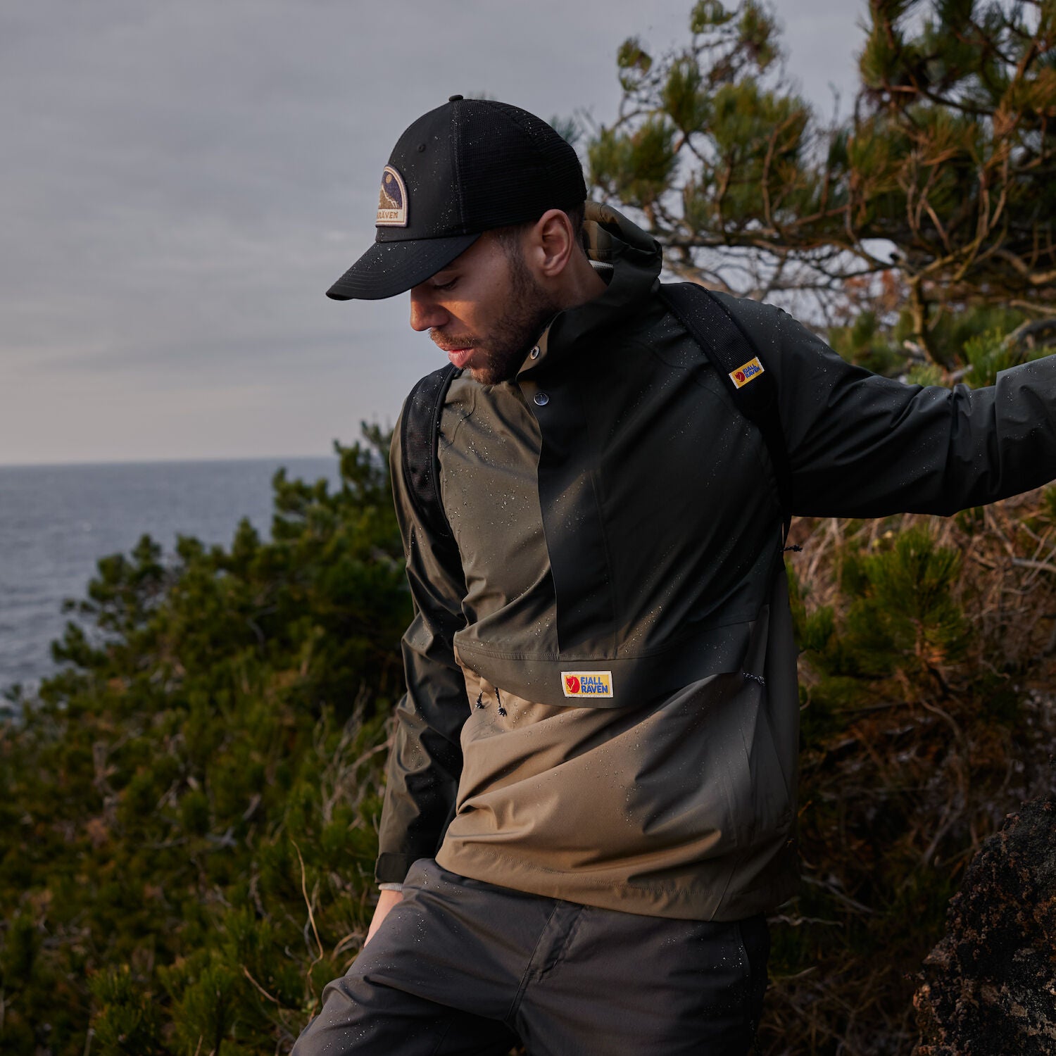 man wearing deep forest-laurel green raincoat jacket with Fjällräven logoand a black cap walking along the forest