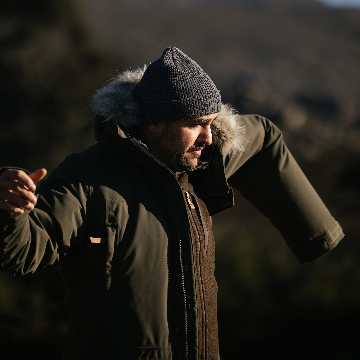 man trying to wear his green parka with fur hood and a grey beanie