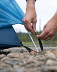 Person setting up a blue tent on a rocky ground with a blurred natural background