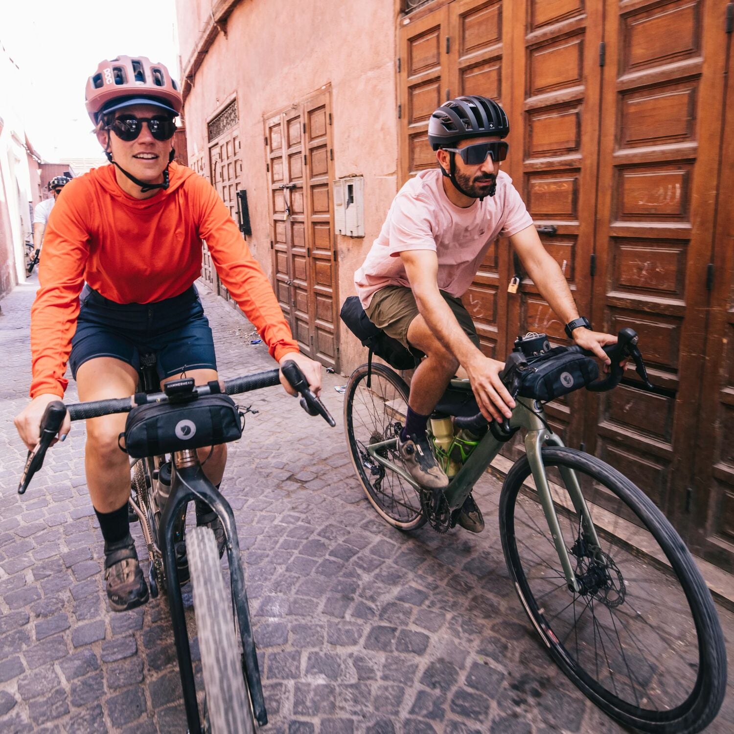 Two cyclists riding bicycles on a cobblestone street with wooden doors in the background.