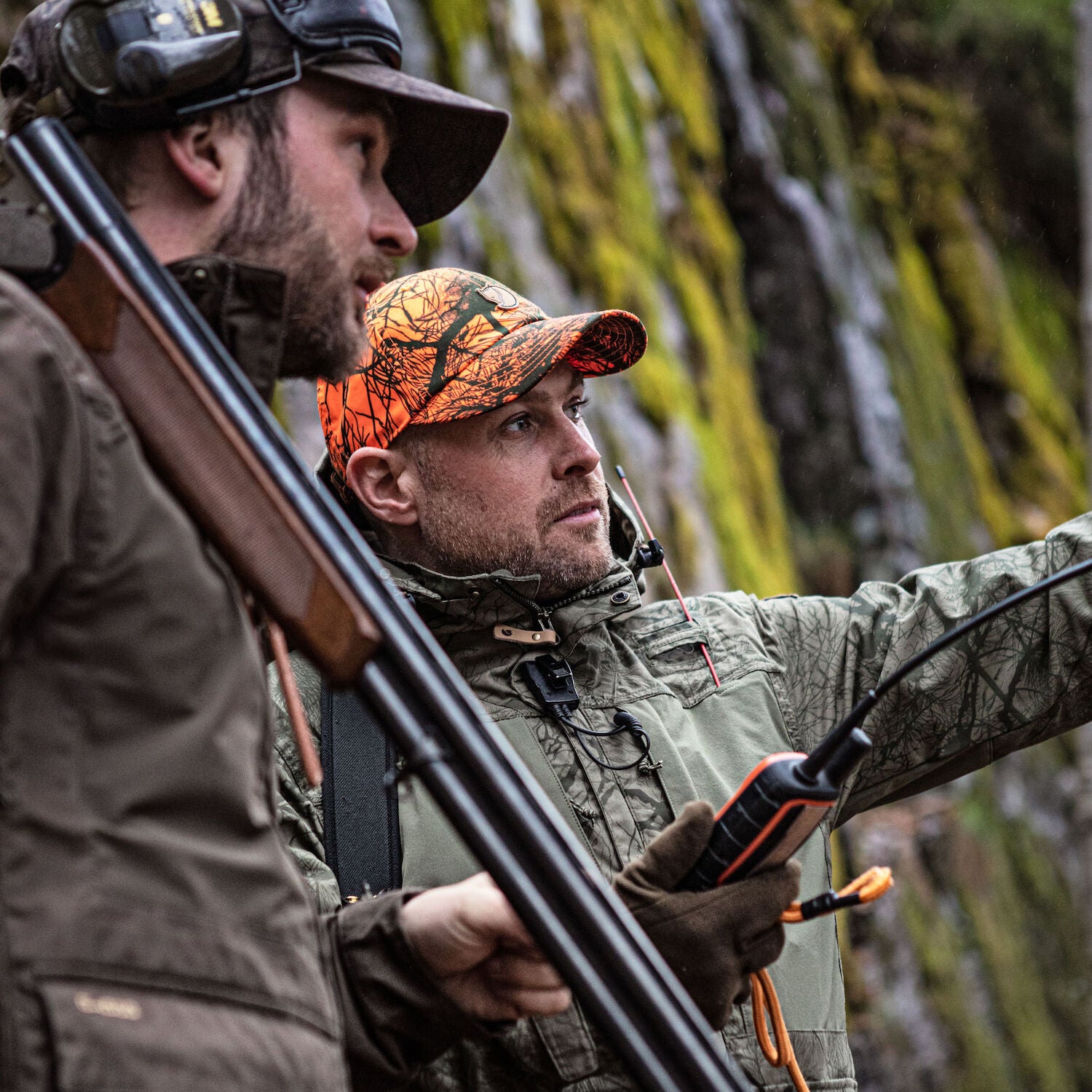 Two hunters in camouflage gear with rifles and communication devices in a forest setting.