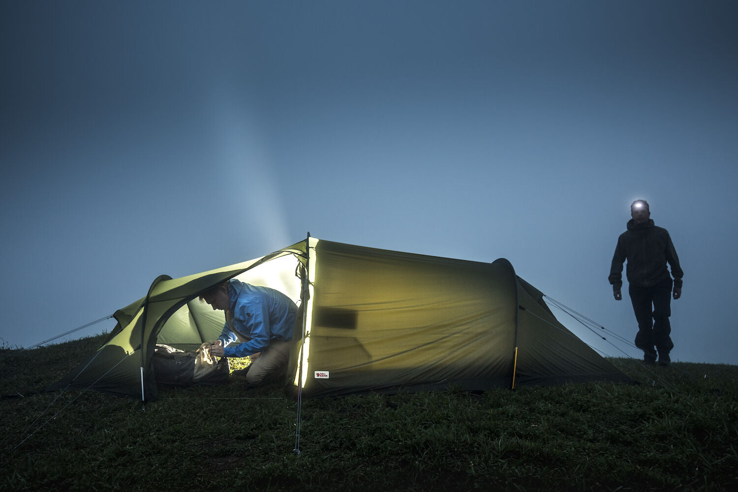 Person inside a tent with a flashlight and another person outside on a grassy hill at night.
