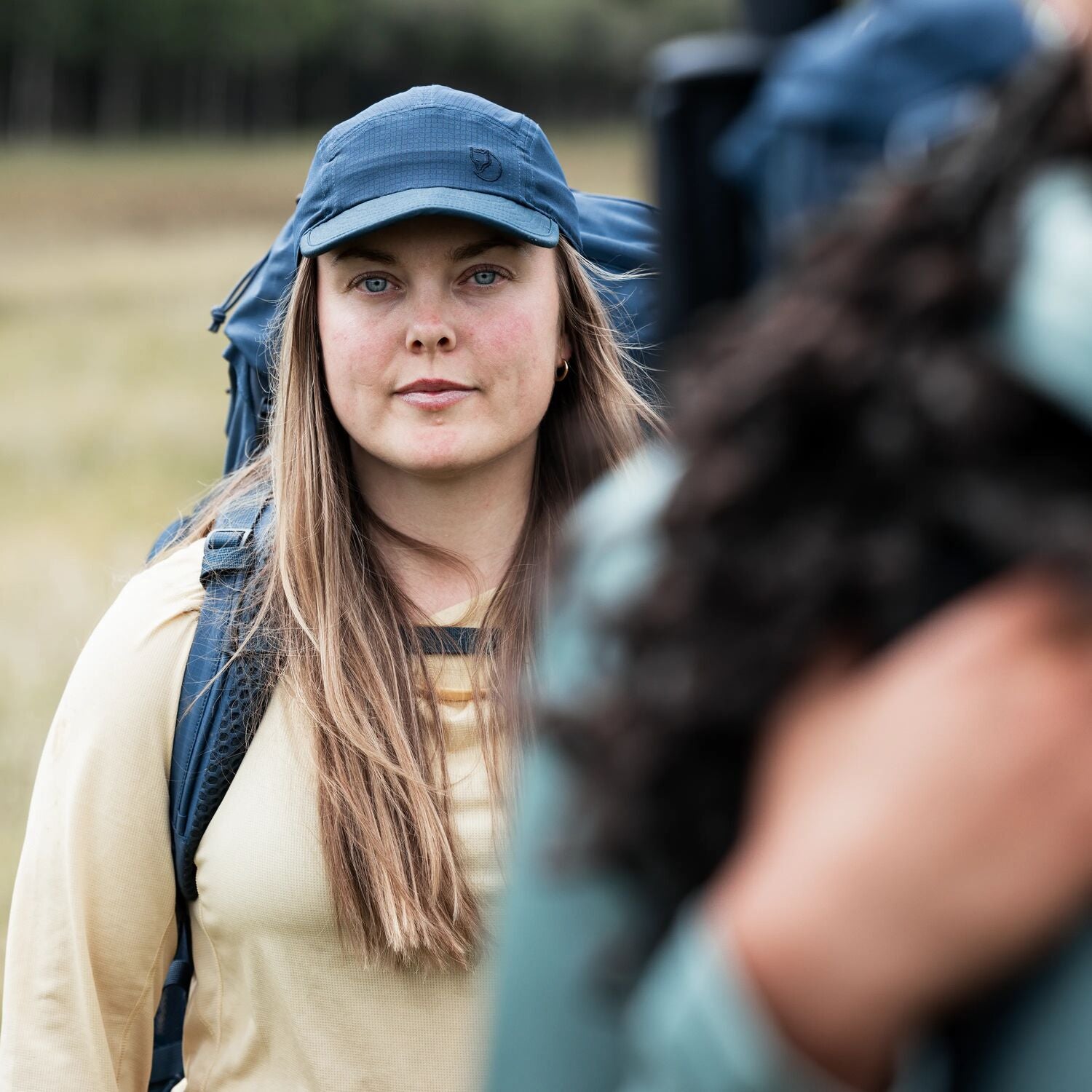 Woman with a blue cap and backpack looking at a camera, with a blurred foreground
