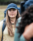 Woman with a blue cap and backpack looking at a camera, with a blurred foreground