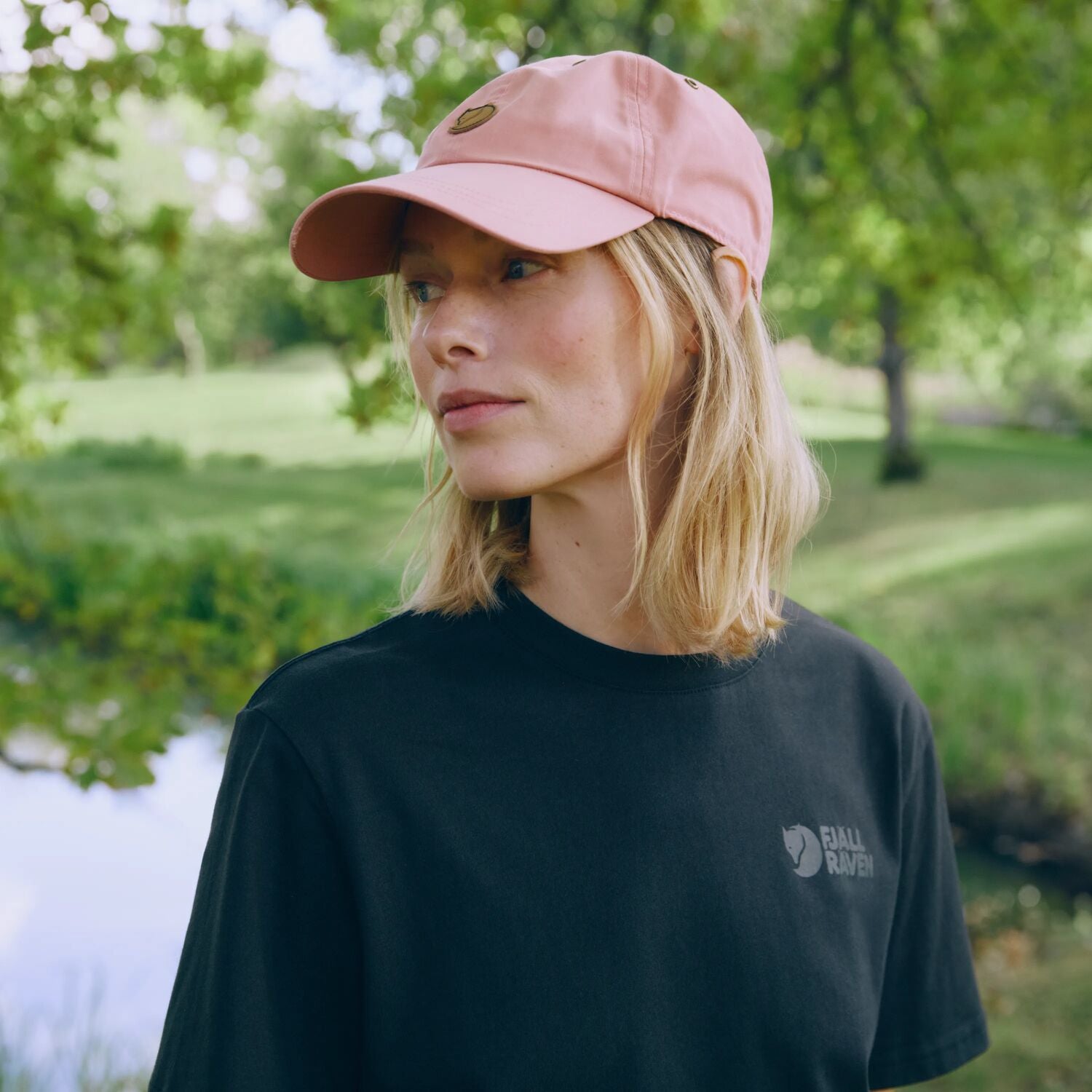 Person wearing a pink cap and black t-shirt with a Fjällräven  logo, standing outdoors.