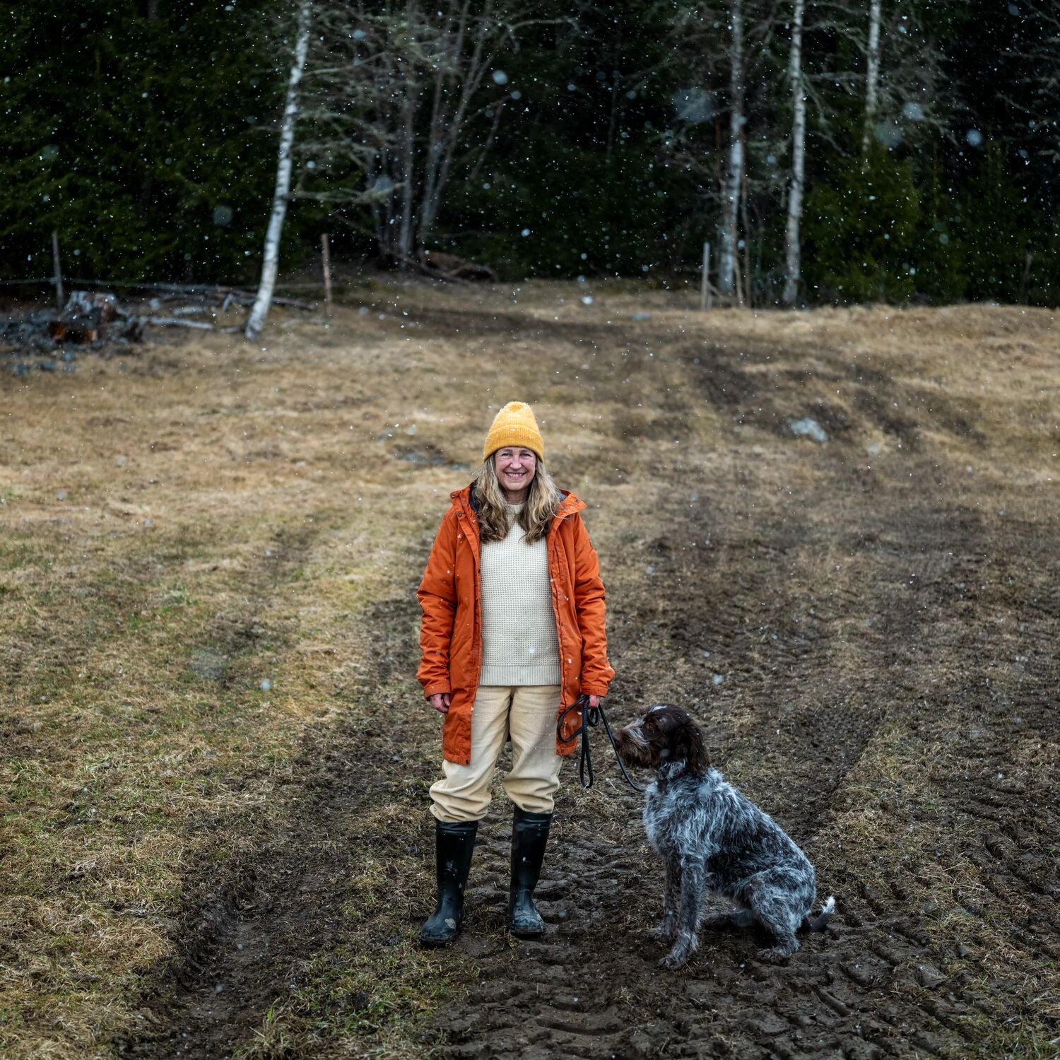 Person in orange jacket and yellow beanie standing with a dog in a forested area