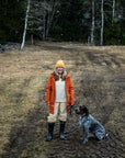Person in orange jacket and yellow beanie standing with a dog in a forested area