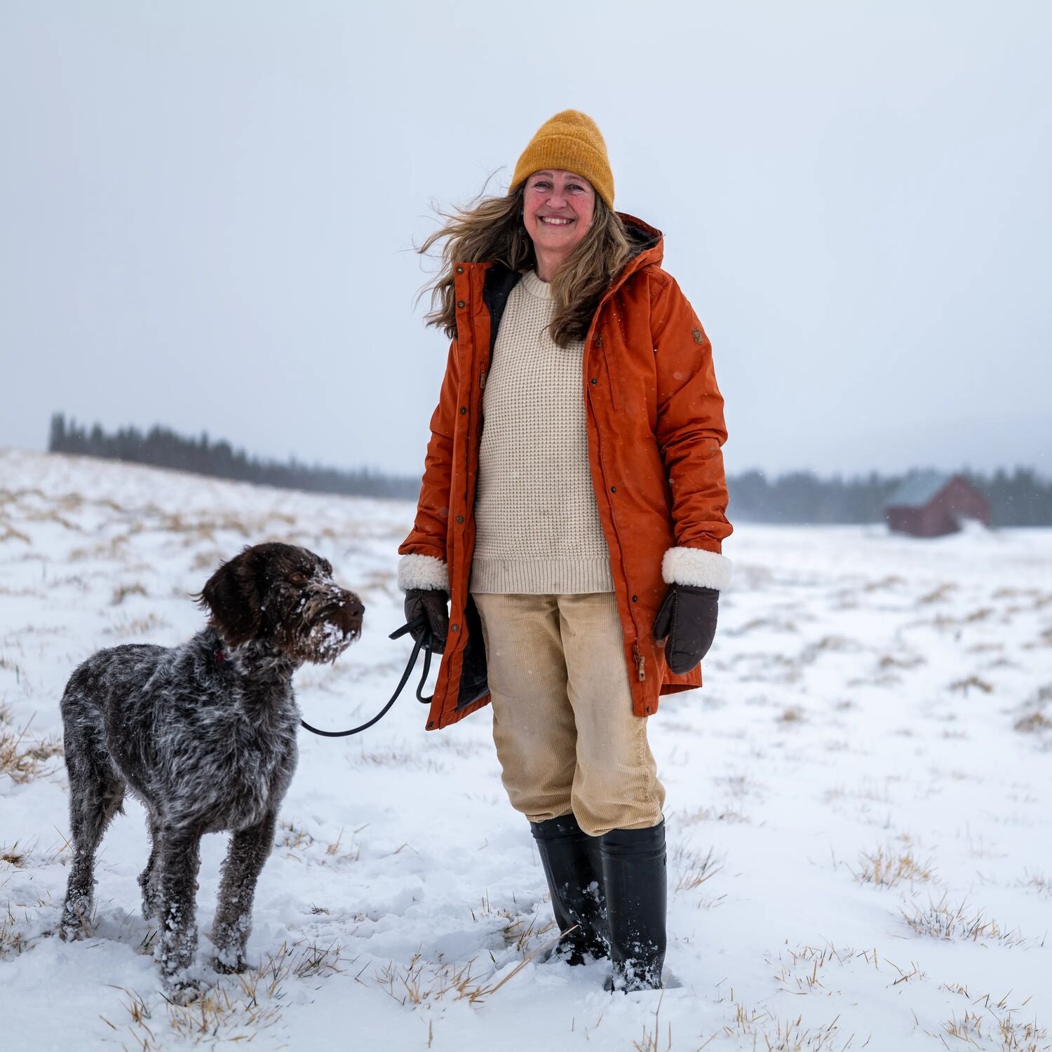 Woman in an orange coat and yellow beanie standing in a snowy field with a dog.