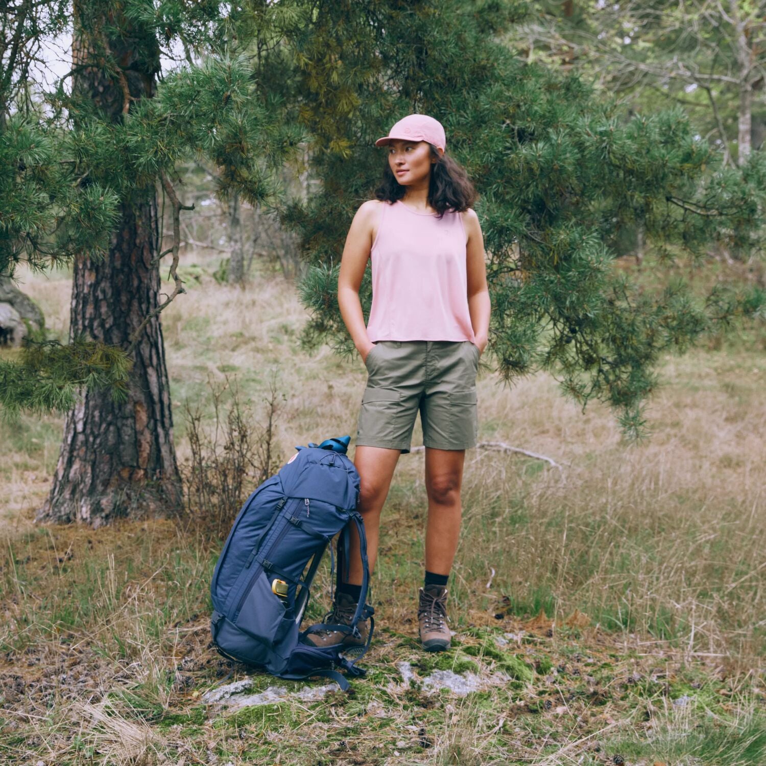 Woman standing in a forest with a blue backpack, wearing a pink sleeveless top and green shorts.