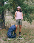 Woman standing in a forest with a blue backpack, wearing a pink sleeveless top and green shorts.