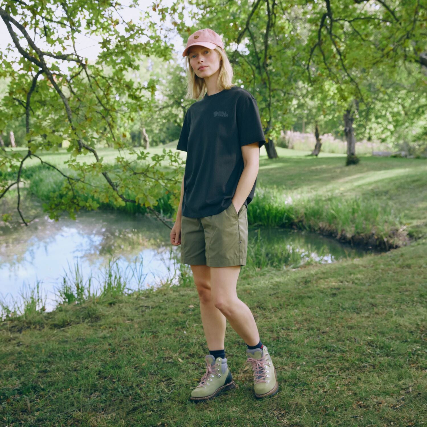 Person standing in a park by a pond wearing a black t-shirt, green shorts, and a pink cap.