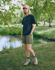 Person standing in a park by a pond wearing a black t-shirt, green shorts, and a pink cap.