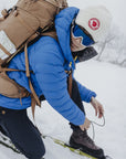 Person preparing to ski in a snowy landscape wearing a blue jacket, brown backpack, and white beanie with a red logo.