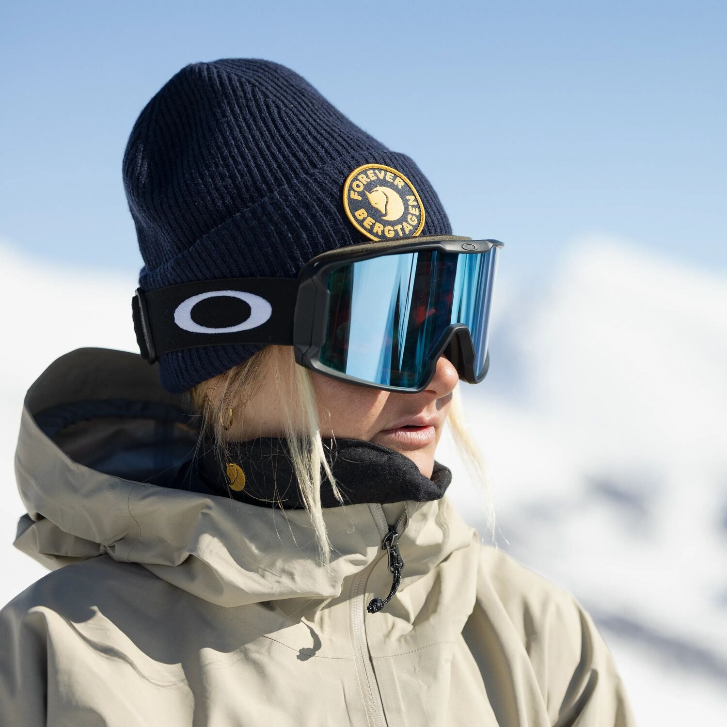 Person wearing a navy beanie with a Fjällräven logo, reflective goggles, and a beige jacket against a snowy background.