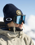 Person wearing a navy beanie with a Fjällräven logo, reflective goggles, and a beige jacket against a snowy background.