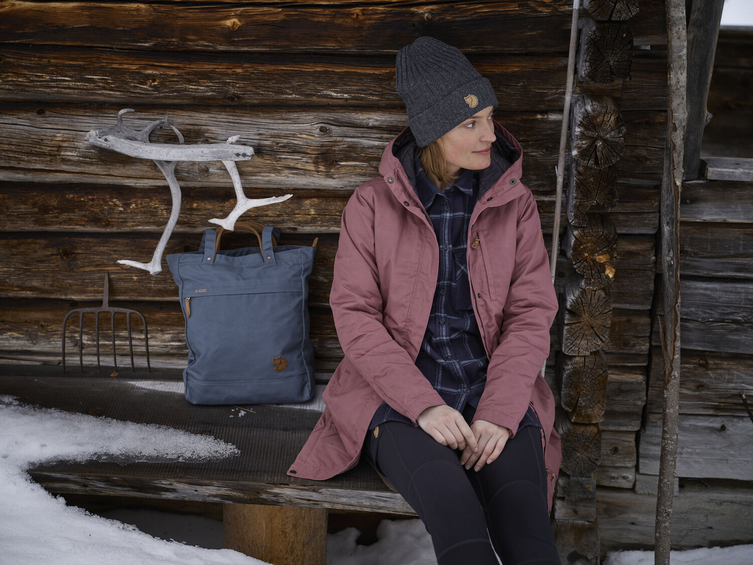 Person wearing a pink coat sitting in front of a wooden cabin with a blue bag beside them.